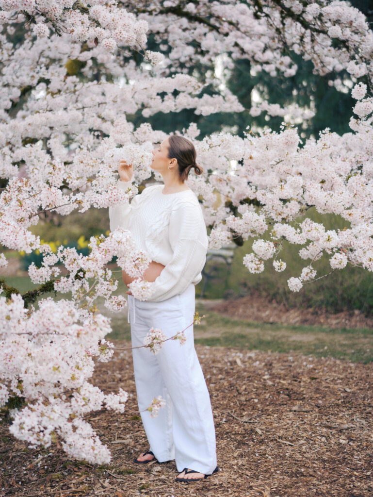 A pregnant woman dressed in white smelling cherry blossom flowers and thinking about the best options for midwives in Seattle 