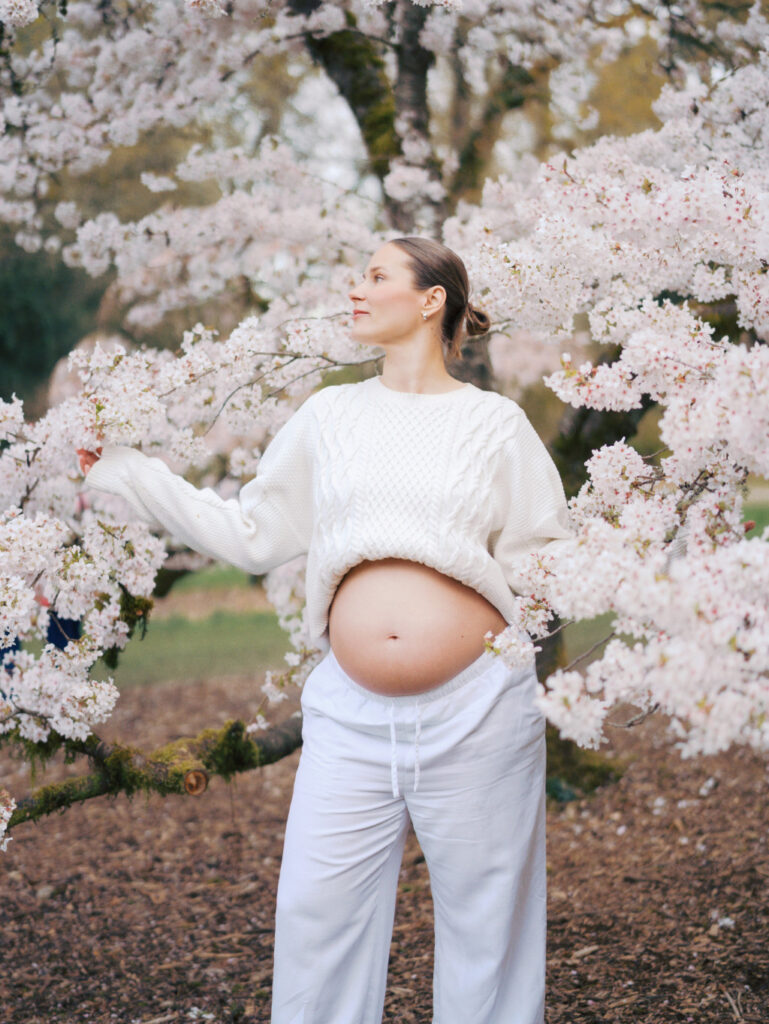 A pregnant mom wearing white is surrounded by cherry blossom trees considering her options for the best midwives in Seattle
