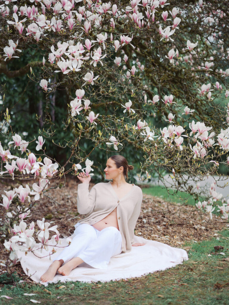 A pregnant woman dressed in white sitting on a blanket under a tree smelling magnolia flowers
