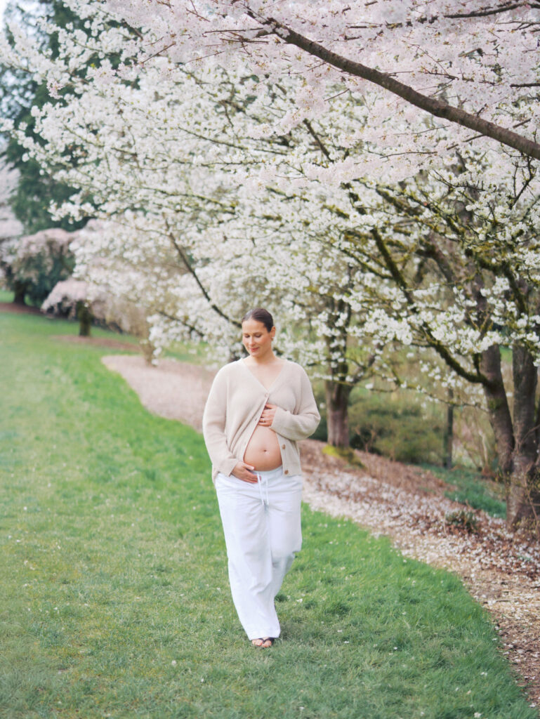 Pregnant woman walking in a cherry blossom garden in Seattle with her belly out wearing an open cardigan