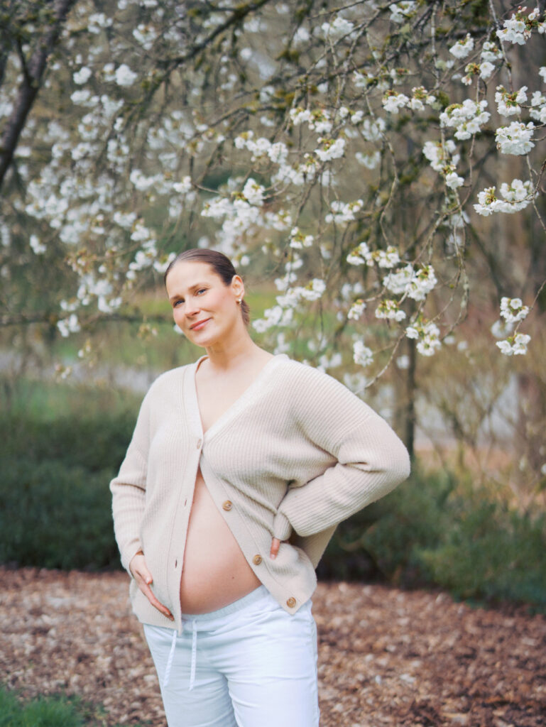 Pregnant woman wearing an open cardigan with her belly out standing casually surrounded by cherry blossoms in a Seattle park