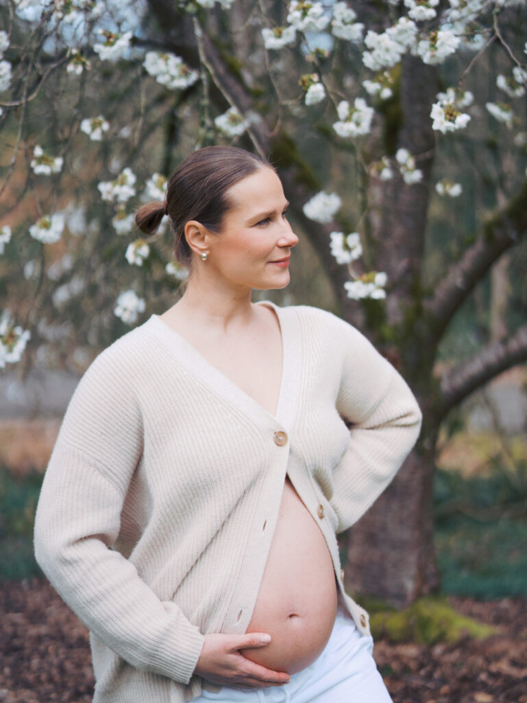 Pregnant woman with her belly out surrounded by cherry blossoms at Washington Park Arboretum standing and looking in the distance