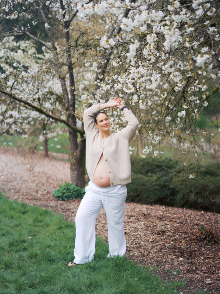 Pregnant woman standing in a cherry blossom garden in Seattle with her arms up 