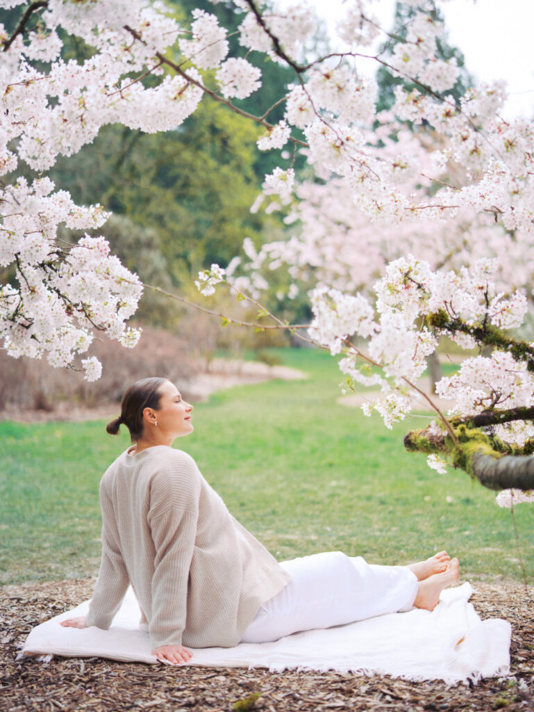 Pregnant woman is facing away and sitting casually on the blanket during cherry blossom season