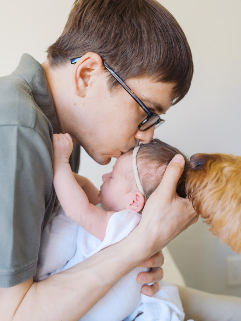 Dad is kissing his newborn baby girl while the dog is sniffing baby's head