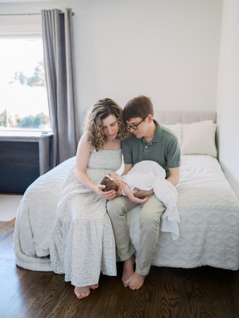Mom and dad sitting next to each other on the bed cuddling their newborn baby girl