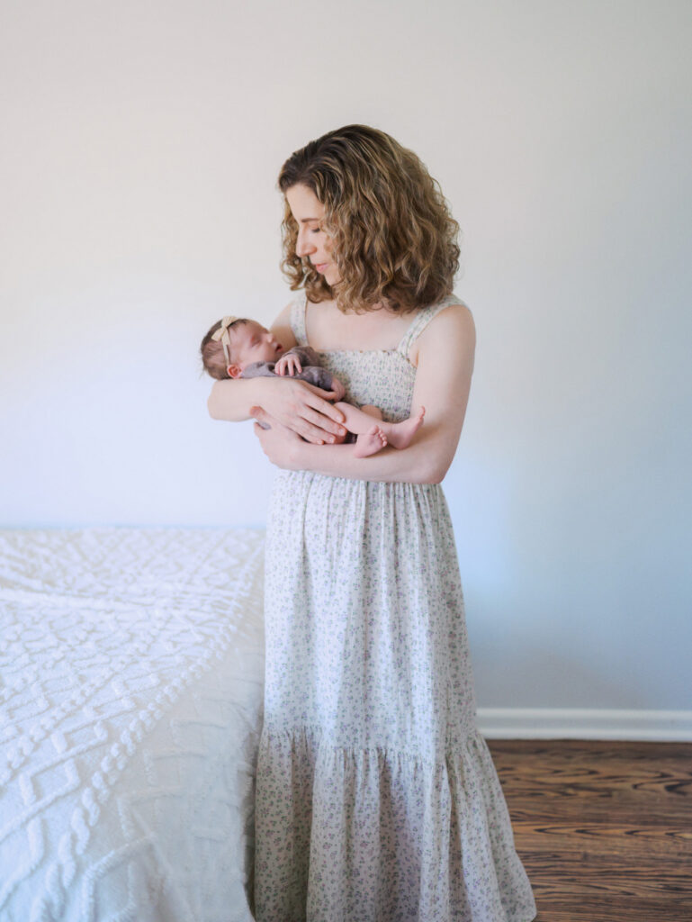 Mom with curly hair wearing a long flowy dress holding her baby girl next to a bed