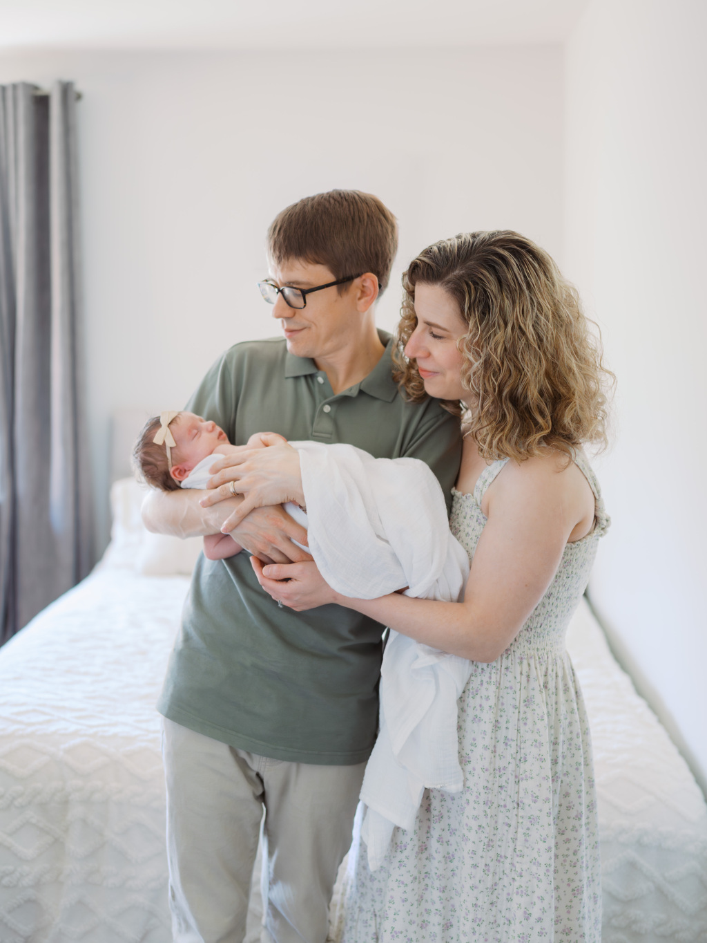 Mom and Dad standing in their bedroom, holding their newborn baby girl wrapped in a swaddle during an in-home baby photography session in Seattle