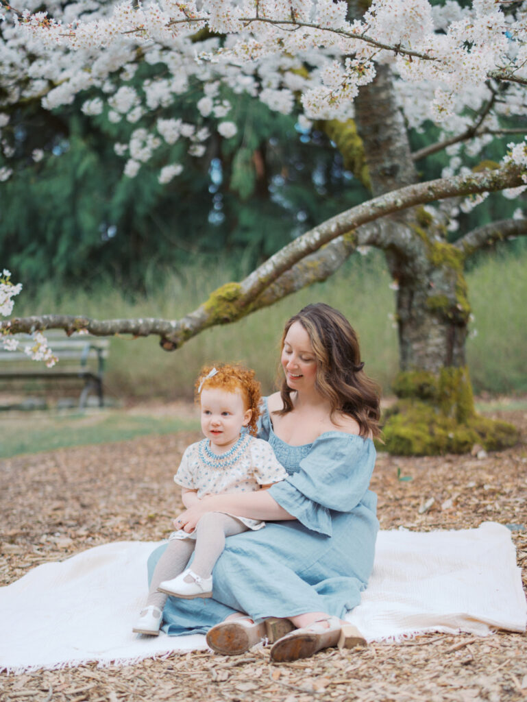 Pregnant mom and her toddler daughter sitting on a blanket in the park surrounded by cherry blossoms