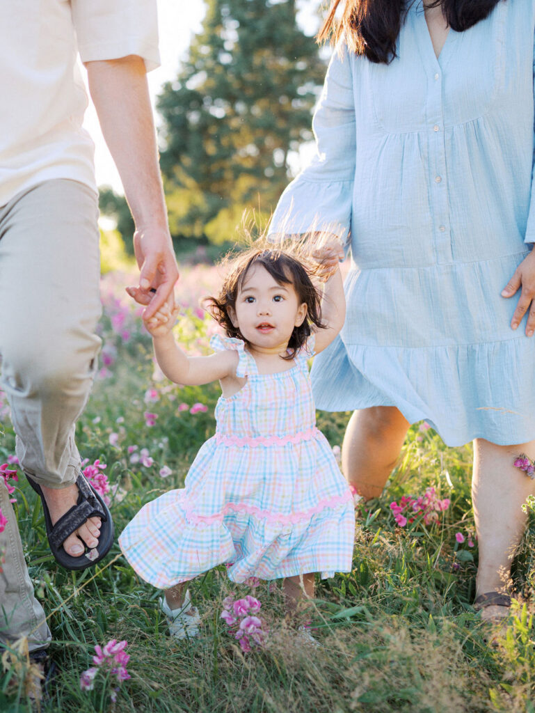 Little girl walking in a flower field with her parents holding her hands